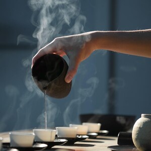 May include: A hand pours hot liquid from a dark brown ceramic vessel into a row of small white teacups. Steam rises from the liquid and cups, suggesting a tea ceremony. The scene is set on a wooden surface with a soft, diffused light.