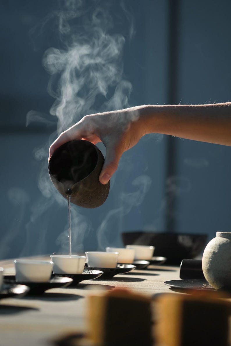May include: A hand pours hot liquid from a dark brown ceramic vessel into a row of small white teacups. Steam rises from the liquid and cups, suggesting a tea ceremony. The scene is set on a wooden surface with a soft, diffused light.