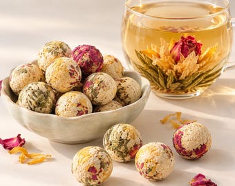 May include: A light-colored ceramic bowl filled with spherical tea balls containing dried flower petals and leaves, alongside a clear glass cup containing a blooming tea flower submerged in golden liquid, with loose petals scattered nearby.