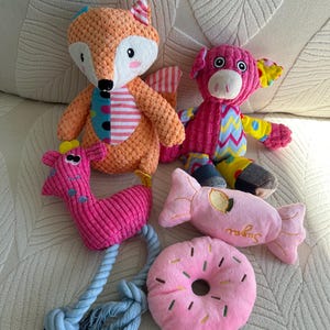 May include: A collection of colorful plush dog toys, including a fox, pig, giraffe, candy, and donut. The fox is orange and white, the pig is pink, and the giraffe is pink with a blue rope. The candy and donut are pink with embroidered details.