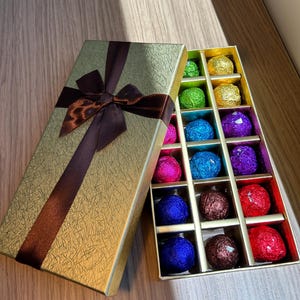 May include: A gold gift box with a brown ribbon and bow, open to reveal a tray of colorful chocolate truffles. The truffles are wrapped in foil of various colors, including green, blue, purple, and red.
