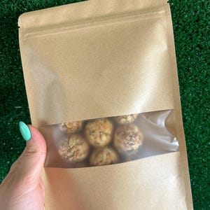 May include: A brown paper bag with a clear plastic window, showing a variety of round, brown, and beige treats inside.