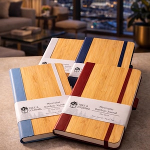 May include: Three rectangular journals with light brown bamboo covers and colored elastic bands in blue, red, and dark blue. Each journal features a white paper belly band printed with the text Tree & Journal, Minimalist Bamboo Journal, and Dot Grid.