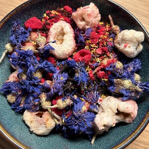 May include: A teal ceramic bowl filled with a colorful mix of ingredients. The mix includes dried blue cornflowers, red raspberries, and white, airy pieces. The ingredients are arranged in a visually appealing way, suggesting a tea blend or a similar product.