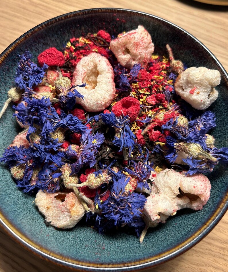 May include: A teal ceramic bowl filled with a colorful mix of ingredients. The mix includes dried blue cornflowers, red raspberries, and white, airy pieces. The ingredients are arranged in a visually appealing way, suggesting a tea blend or a similar product.