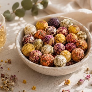 May include: A speckled ceramic bowl filled with numerous round, compressed tea balls made of dried flower petals and herbs in shades of yellow, pink, purple, and green, accompanied by a glass cup containing a light golden herbal tea infusion.