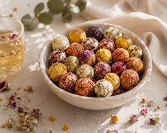 May include: A speckled ceramic bowl filled with numerous round, compressed tea balls made of dried flower petals and herbs in shades of yellow, pink, purple, and green, accompanied by a glass cup containing a light golden herbal tea infusion.