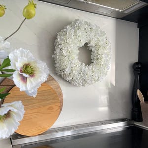 May include: A white faux flower wreath hangs on a white kitchen backsplash. The wreath is made of white hydrangeas and is about 30 cm in diameter. There is a wooden chopping board and a white flower on the counter below the wreath.