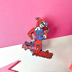 Puede incluir: Un pin de esmalte rojo, azul y negro con un personaje de dibujos animados con un gorro de bufón y el texto "Daddy's Lil' Nymphomaniac".