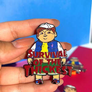 K&ouml;nnte beinhalten: Eine rote Emaille-Anstecknadel mit einer Comicfigur, die eine blaue Jacke, ein gelbes Hemd und eine gr&uuml;ne Shorts tr&auml;gt. Die Figur steht mit dem Text "SURVIVAL OF THE THICKEST" in Rot.