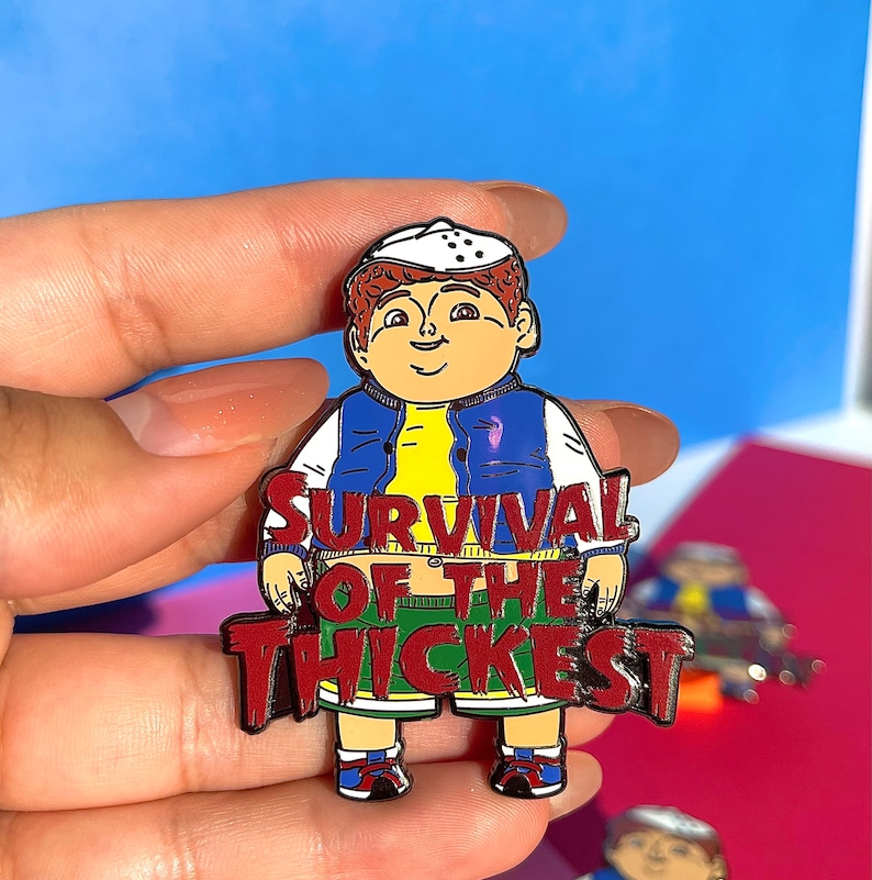 K&ouml;nnte beinhalten: Eine rote Emaille-Anstecknadel mit einer Comicfigur, die eine blaue Jacke, ein gelbes Hemd und eine gr&uuml;ne Shorts tr&auml;gt. Die Figur steht mit dem Text "SURVIVAL OF THE THICKEST" in Rot.