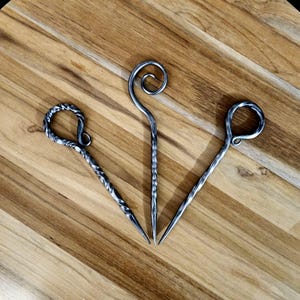 May include: Three metal skewers with twisted handles and decorative loops at the top. The skewers are arranged in a triangular shape on a wooden surface.