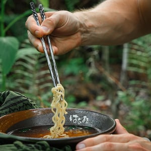 Op de afbeelding: Een paar gestructureerde, donker metalen eetstokjes die een portie lichtgekleurde noedels uit een donkere, ronde keramische kom met bouillon tillen. De kom heeft een gespikkelde afwerking en witte Japanse tekens op de buitenrand.