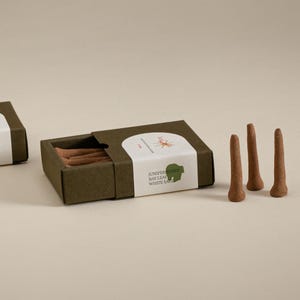 May include: A box of incense sticks with a white label that reads "Juniper Berry, Bay Leaf, White Sage." The box is open, revealing several brown incense sticks. Three additional incense sticks stand upright next to the box.