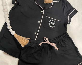 May include: Black pajama set with short sleeves and shorts. The top has a white piping trim and a button-up front. The shorts have a pink drawstring and white piping trim. The top has a pocket with a monogrammed "LGS" in white.