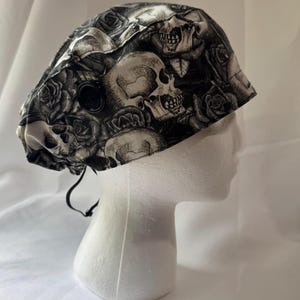 May include: A black and white skull and rose patterned surgical scrub cap. The cap is made of fabric and features a button and adjustable ties. The design includes multiple skulls and roses.