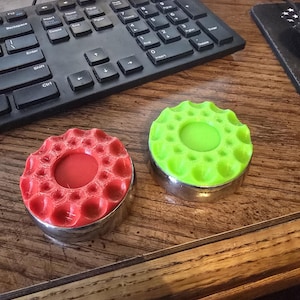 May include: Two round, decorative objects with a textured, dimpled design. One is red, the other is green, both with a silver metal base. The objects are on a wooden surface near a black keyboard.