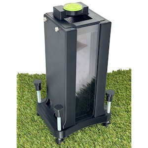 May include: A black rectangular device with a clear front panel, resting on a green surface. The device has a green circular button on top and adjustable feet. The device appears to be a container or dispenser.