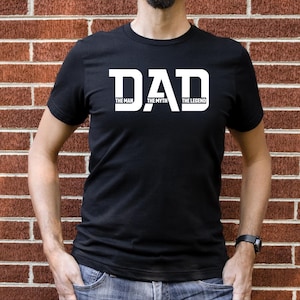 May include: Black t-shirt with the word "DAD" in large white capital letters. Below the word "DAD" are the words "THE MAN", "THE MYTH", and "THE LEGEND" in smaller white capital letters. The person is wearing blue jeans.