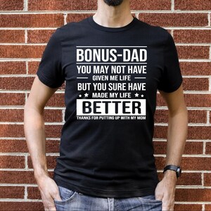 May include: Black t-shirt with white text that reads "Bonus-Dad You may not have given me life but you sure have made my life better Thanks for putting up with my mom."