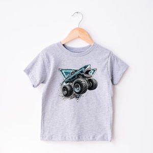 May include: A light grey t-shirt with a graphic of a monster truck with a shark mouth design. The truck is black and teal, with large black tyres. The graphic is set within a teal and white triangle.