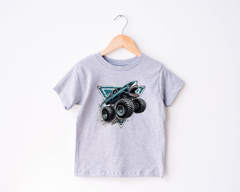 May include: A light grey t-shirt with a graphic of a monster truck with a shark mouth design. The truck is black and teal, with large black tyres. The graphic is set within a teal and white triangle.