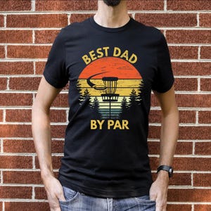 May include: A black t-shirt with a vintage-style graphic of a disc golf basket in front of a sunset. The text "Best Dad By Par" is printed on the shirt.