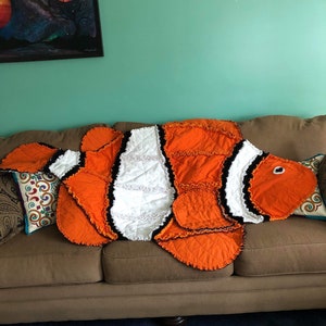 May include: A handmade orange, white, and black patchwork quilt in the shape of a clownfish. The quilt is lying on a brown couch.