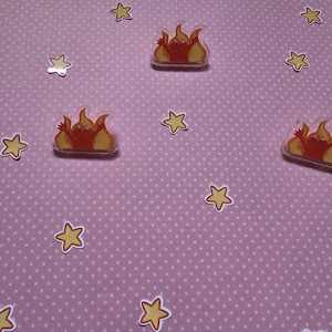May include: A collection of stickers featuring a cartoon flame design in shades of orange and yellow. The stickers are scattered across a pink surface with white polka dots, along with star-shaped stickers.