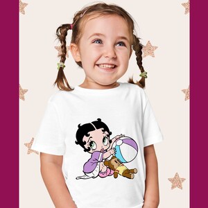 May include: A white t-shirt featuring a cartoon image of a girl with black hair and a purple dress playing with a beach ball and a teddy bear.