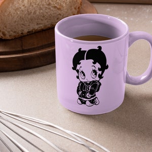 May include: A lavender ceramic mug featuring a black and white illustration of Betty Boop wearing a jacket with a letter "B" on it. The mug is filled with coffee.