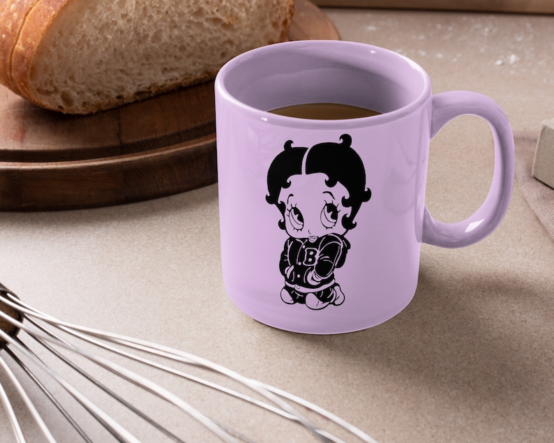 May include: A lavender ceramic mug featuring a black and white illustration of Betty Boop wearing a jacket with a letter "B" on it. The mug is filled with coffee.