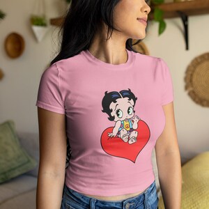 May include: A pink cropped t-shirt featuring a cartoon image of a baby girl sitting on a red heart. The baby girl is wearing a blue and yellow outfit and has black hair and large blue eyes.