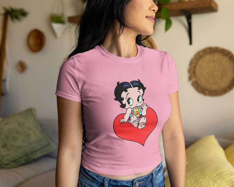 May include: A pink cropped t-shirt featuring a cartoon image of a baby girl sitting on a red heart. The baby girl is wearing a blue and yellow outfit and has black hair and large blue eyes.