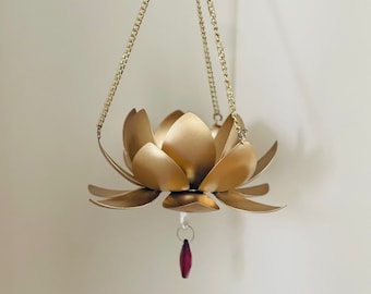 May include: A gold-coloured metal lotus flower hanging ornament. The flower has multiple petals and is suspended by a gold chain. A small, red crystal hangs from the bottom of the flower.
