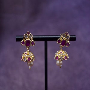 Op de afbeelding: Een paar gouden oorbellen met roze en witte stenen. De oorbellen hangen aan een zwarte standaard.