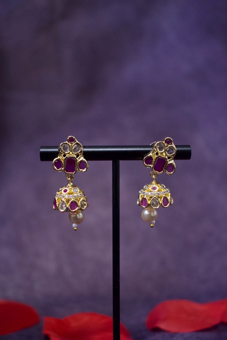 Op de afbeelding: Een paar gouden oorbellen met roze en witte stenen. De oorbellen hangen aan een zwarte standaard.
