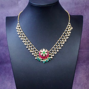 Op de afbeelding: Een gouden ketting met een hanger met een rood en groen bloemmotief. De ketting is gemaakt van kleine witte parels en heeft een delicate ketting.