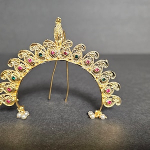 May include: A gold-colored hair ornament with a decorative design featuring green and pink gemstones. The ornament is shaped like a crescent moon and has two pearl-like accents on each side.