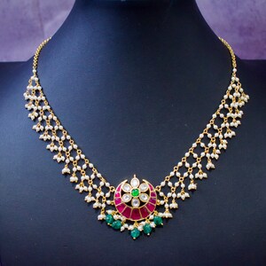 Op de afbeelding: Een gouden ketting met een hanger met een ontwerp van roze en groene edelstenen. De ketting is gemaakt van kleine witte parels en gouden kettingen.