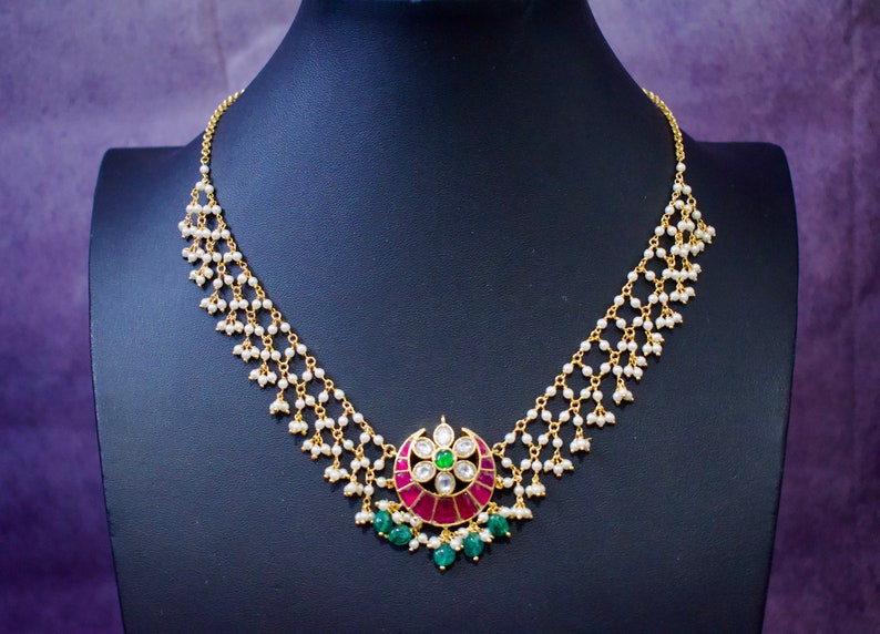 Op de afbeelding: Een gouden ketting met een hanger met een ontwerp van roze en groene edelstenen. De ketting is gemaakt van kleine witte parels en gouden kettingen.