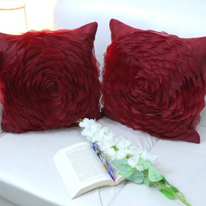 May include: Two burgundy decorative throw pillows with a large, 3D rose design. The pillows are on a white couch with a book and white flowers.