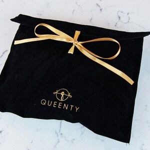 Pu&ograve; includere: Borsa in velluto nero con un nastro dorato legato in un fiocco. La borsa ha il testo "QUEENTY" stampato in oro.
