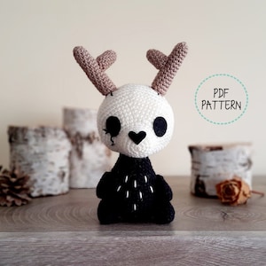 Puede incluir: Patrón de amigurumi de crochet para un pequeño ciervo de calavera blanca con cuernos negros y cuerpo negro. El ciervo tiene una nariz en forma de corazón y ojos negros. El patrón está disponible para descargar en PDF.
