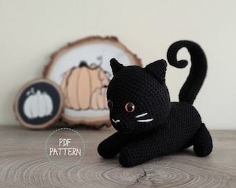 May include: A black crochet cat with orange eyes and white whiskers. The cat is lying down with its tail curled. The image also includes wooden pumpkin decorations and the text "PDF PATTERN".