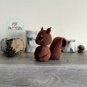 May include: A brown crocheted squirrel amigurumi toy with black eyes and a white nose. The toy is sitting on a wooden surface with a birch log and pine cone in the background.