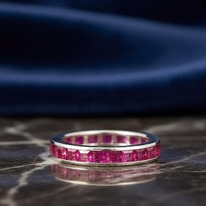 May include: A silver-toned eternity band ring featuring a continuous row of square-cut, vibrant pinkish-red gemstones set in a channel setting. The ring displays a polished metallic finish and a uniform design around the entire circumference.