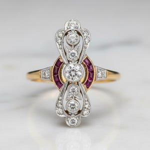 Op de afbeelding: Een ring in vintage-stijl met een gouden band en een centraal ontwerp met een grote, ronde diamant. Het ontwerp wordt geflankeerd door kleinere diamanten en levendige rode edelstenen, wat een opvallend contrast creëert. De ingewikkelde details en het elegante ontwerp van de ring maken het een prachtig sieraad.