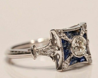 May include: A silver ring with a square, Art Deco-style setting. The setting features a central, round diamond surrounded by blue gemstones and smaller diamonds. The ring has intricate detailing and a polished finish.