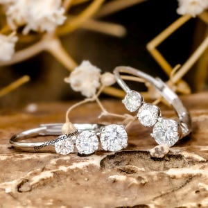 May include: Two silver rings, each set with multiple clear, round gemstones. The rings are displayed on a wooden surface, with white flowers in the background. The text "ReminiscenceJewel" is visible on one of the rings.